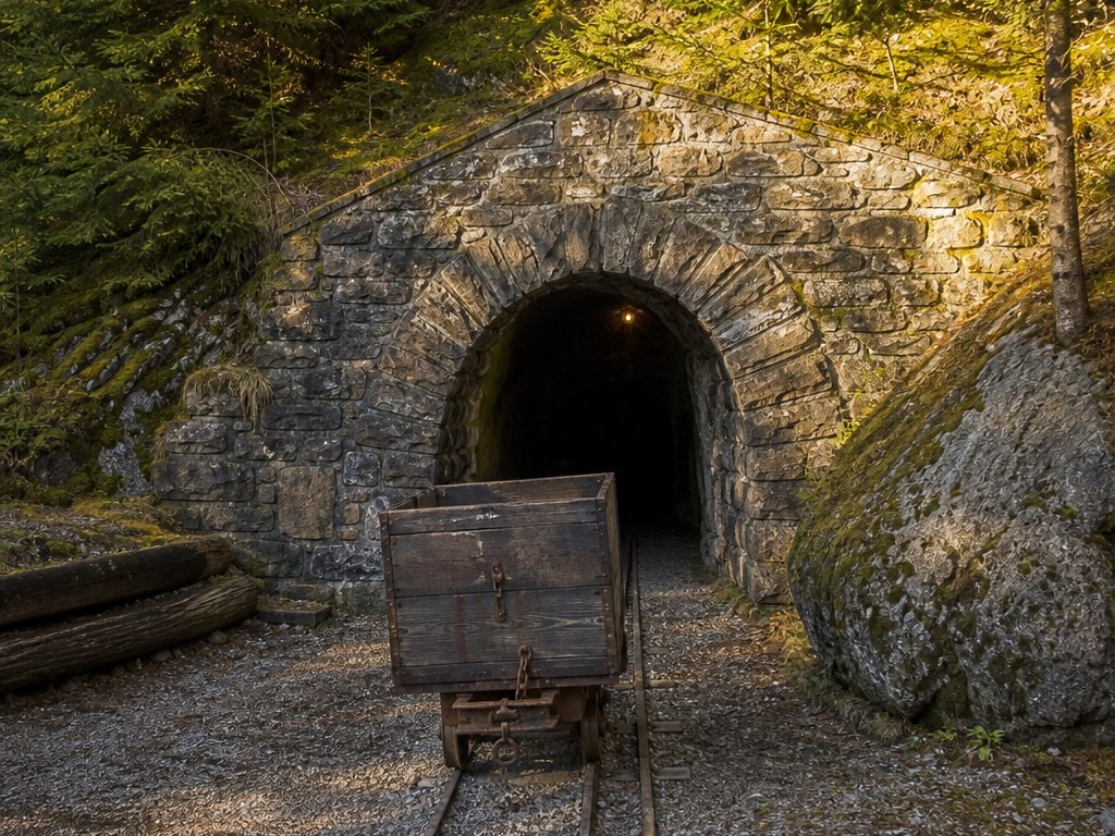 Gemauertes Mundloch eines alten Stollens im bewaldeten Hang, davor Gleise und ein hölzerner Förderwagen (mit AI erstelltes Bild)