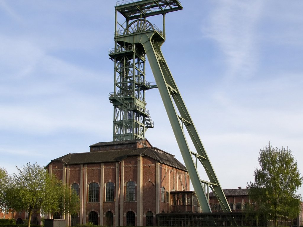 Stählerner Förderturm einer stillgelegten Zeche vor blauem Himmel, davor eine Maschinenhalle aus Backstein (mit AI erstelltes Bild)